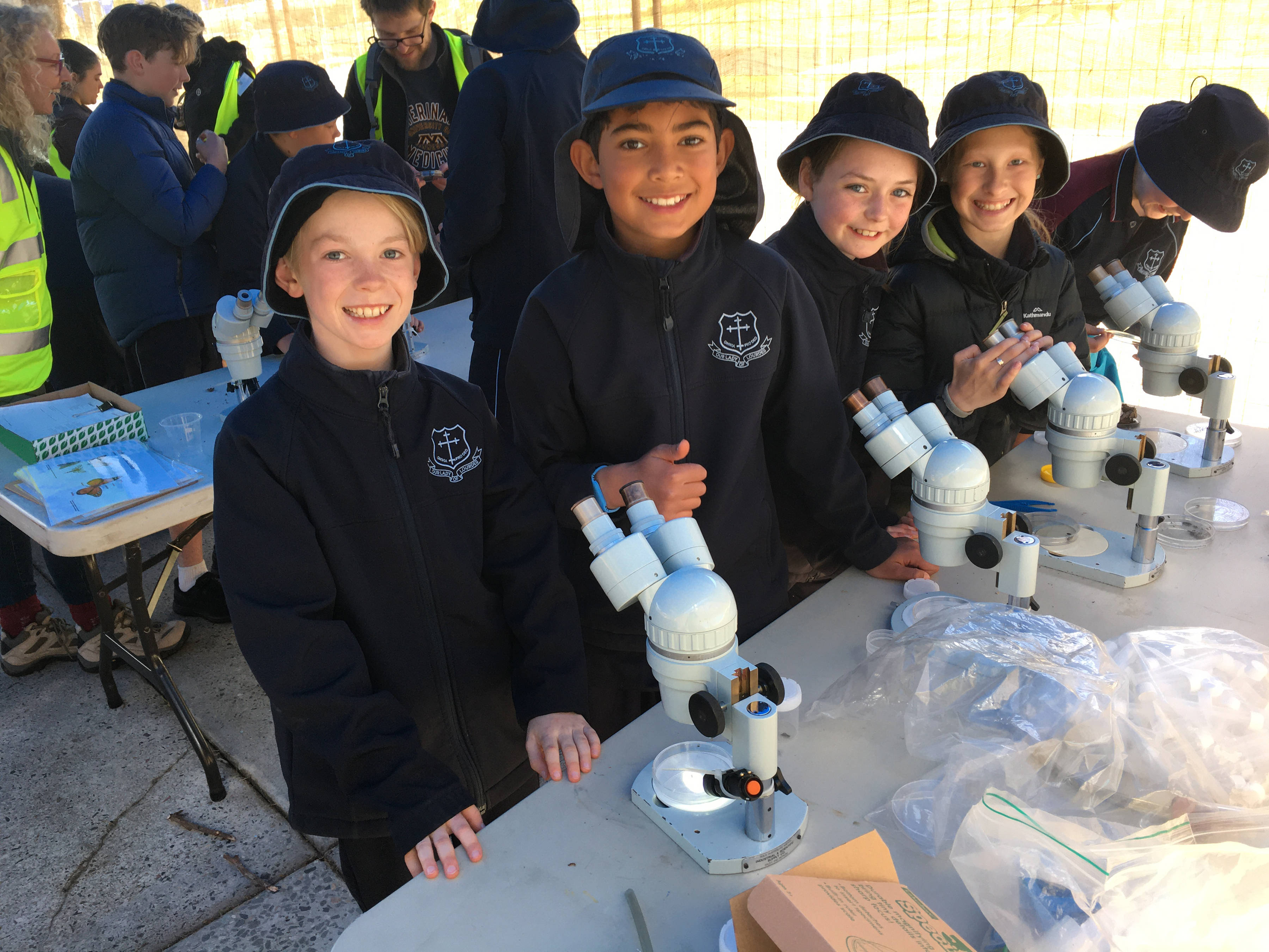 Five primary school students, in navy blue, long sleeved uniforms and hats. Four of the students are looking directly at the camera and smiling, one is busy with a microscope. They are undercover but outside and standing behind a table that has four microscopes on it. There are more students in the background, engaged in other activities.