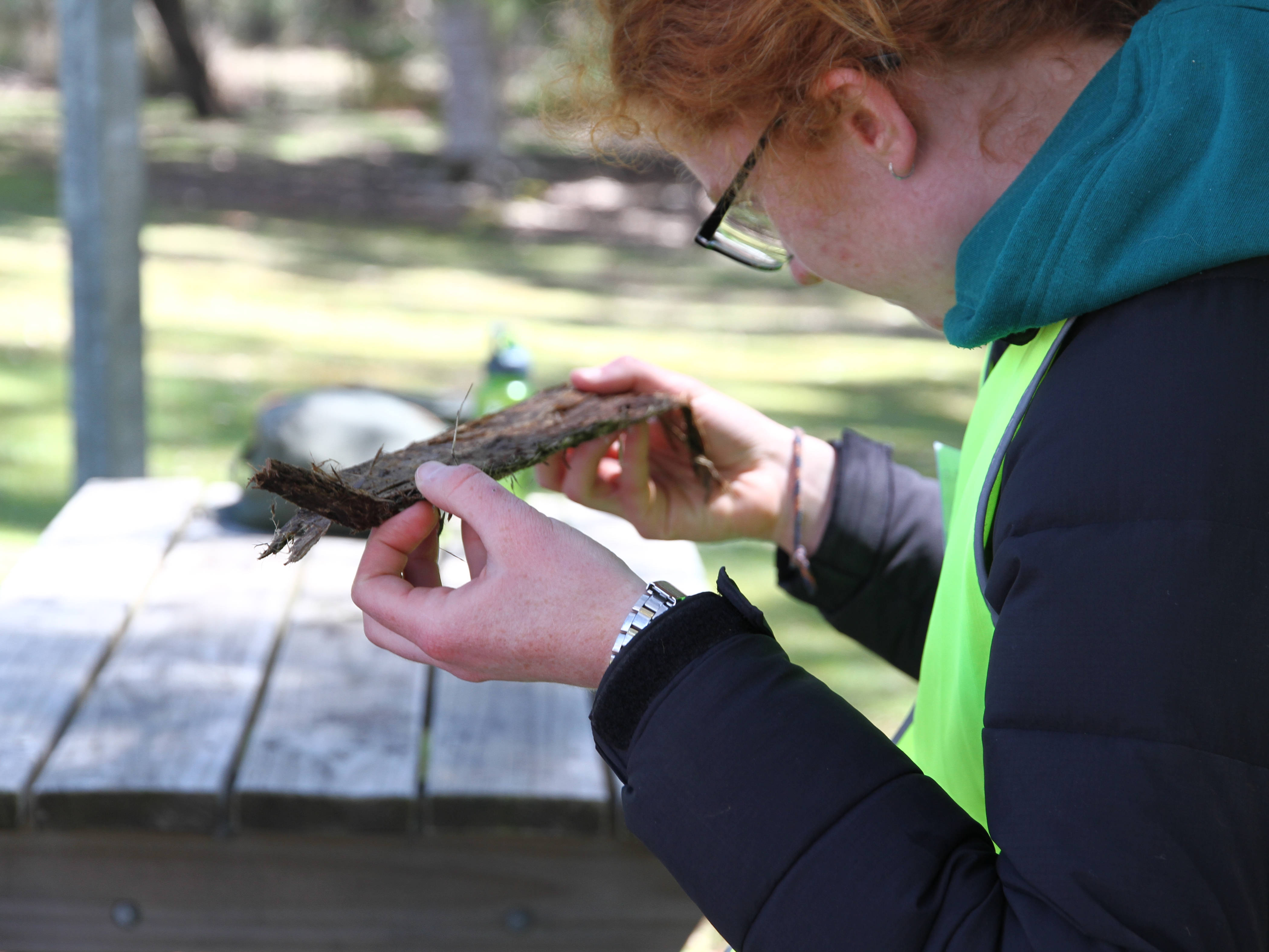School student looking intently at a piece of bark. The student has red hair and glasses and is wearing a hoodie with a green high-vis vest. They are holding a dark brown piece of tree bark.
